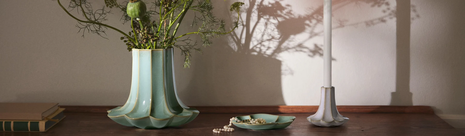 Uurteinen turkoosinvärinen maljakko lehtineen, helmikaulakoru yhteensopivassa astiassa, valkoinen kynttilä uurteisessa pidikkeessä ja kirjoja puupinnalla.