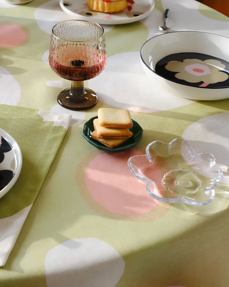 Pöytä katettuna kuvioidulla viinilasilla, neliönmuotoisilla kekseillä vihreällä lautasella, kukkakulholla ja vaaleanvihreällä pöytäliinalla, jossa on vaaleanpunaisia ja valkoisia ympyröitä.