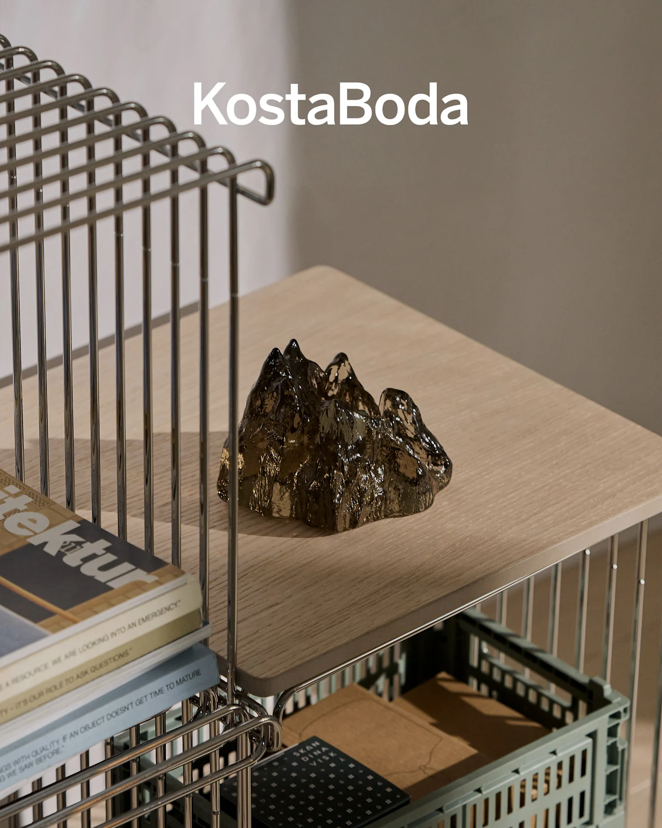 KostaBoda-tuotemerkki tumman, läpinäkyvän, vuorenmuotoisen lasiveistoksen yläpuolella vaalealla puuhyllyllä.