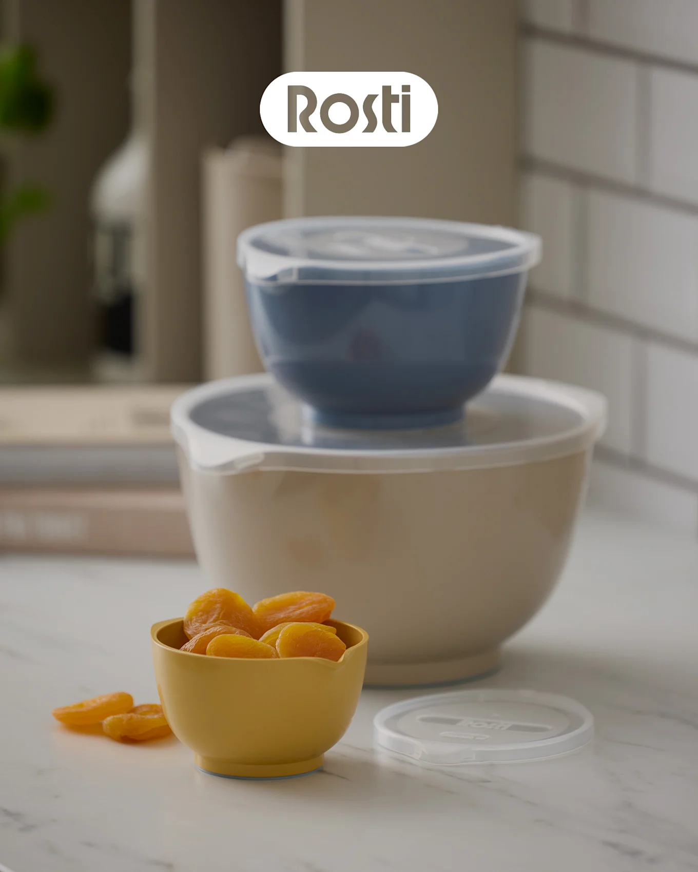 Kolme Rosti-kulhoa tiskillä: beige kulho, jonka päällä on sininen, molemmissa kannet, sekä pieni keltainen kulho täynnä kuivattuja aprikooseja.