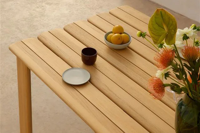 Vaalea puinen lamellipöytä, jossa kulho sitruunoita, tumma muki, pieni lautanen ja maljakko oransseja ja valkoisia kukkia.