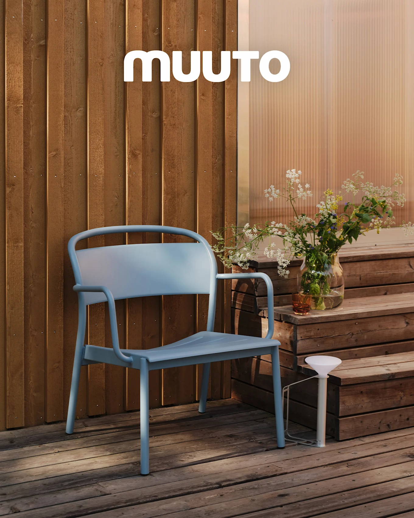 Vaaleansininen ulkotuoli puuterassilla portaiden vieressä, jossa on maljakko luonnonkukkia ja valkoinen kannettava lamppu. MUUTO-logo.