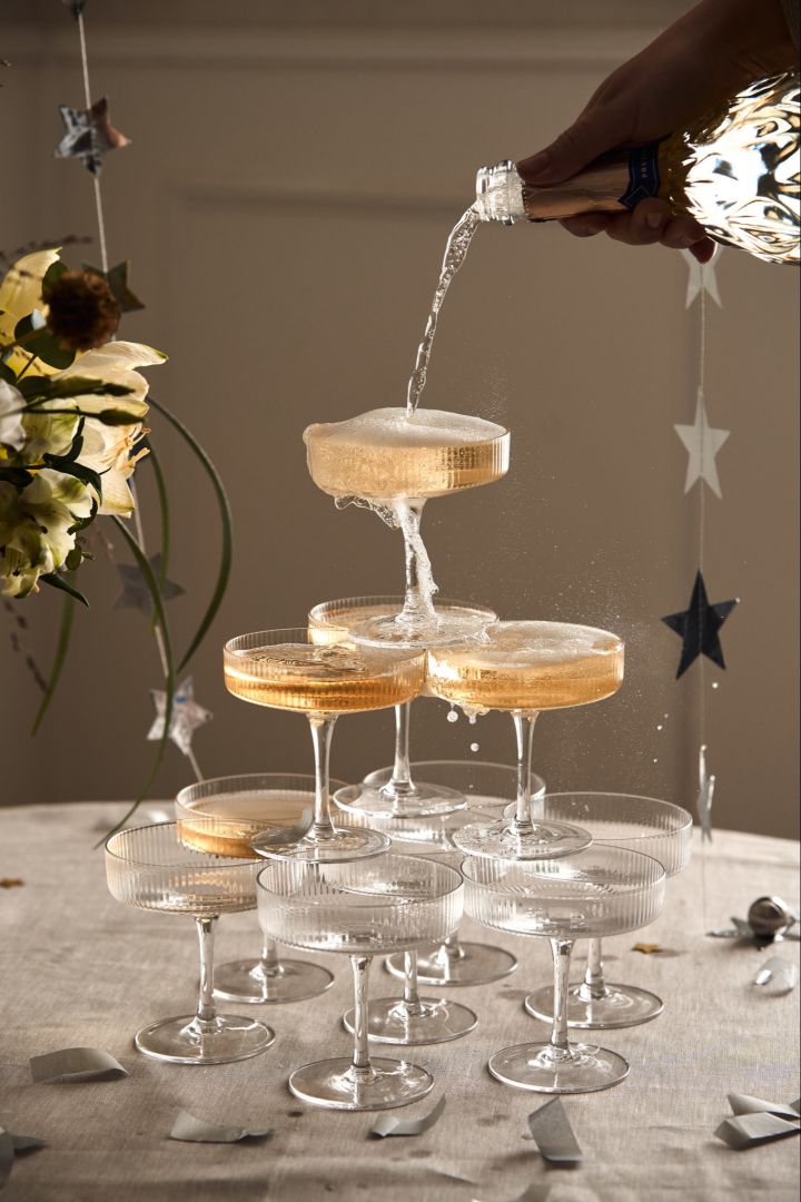 Opi rakentamaan samppanjatorni, kuten tässä kuvassa, jossa käsi kaataa samppanjaa aaltoilevista samppanjalaseista tehdyn pienen samppanjatornin ylimpään lasiin. 