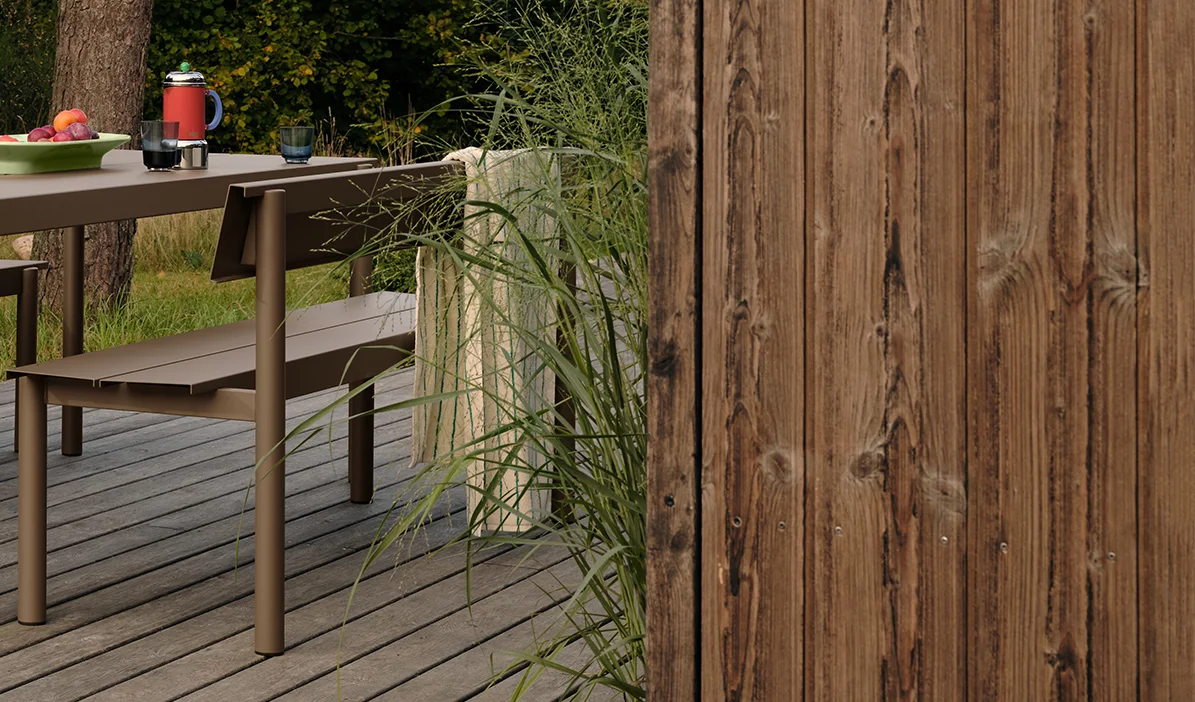 Ruskea metallinen ulkopöytä ja penkki puukannella hedelmien ja juomien kera, korkean ruohon ja puuaidan vieressä.